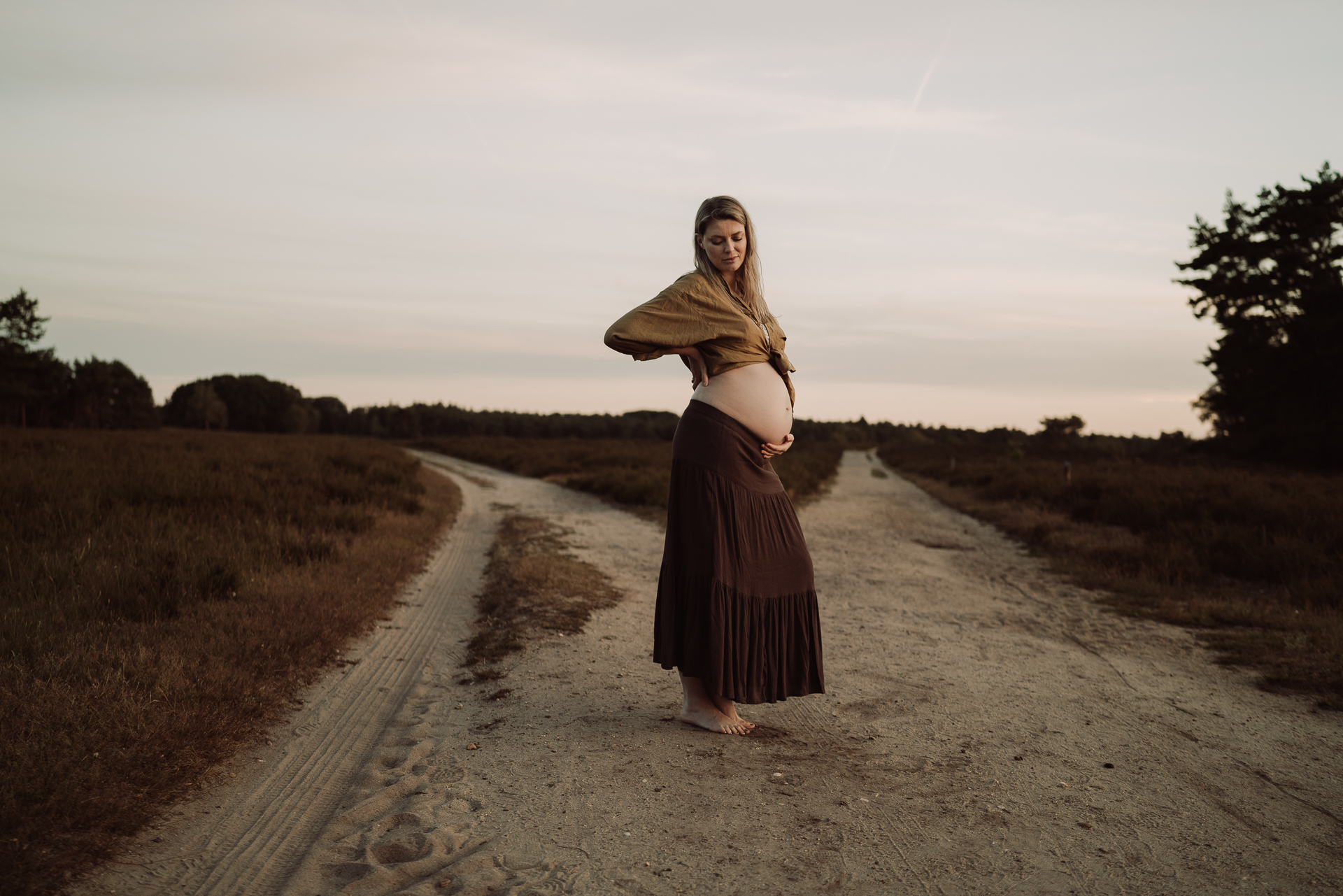 Zwangere vrouw met ontblote buik staat op een zandpad tussen de heide