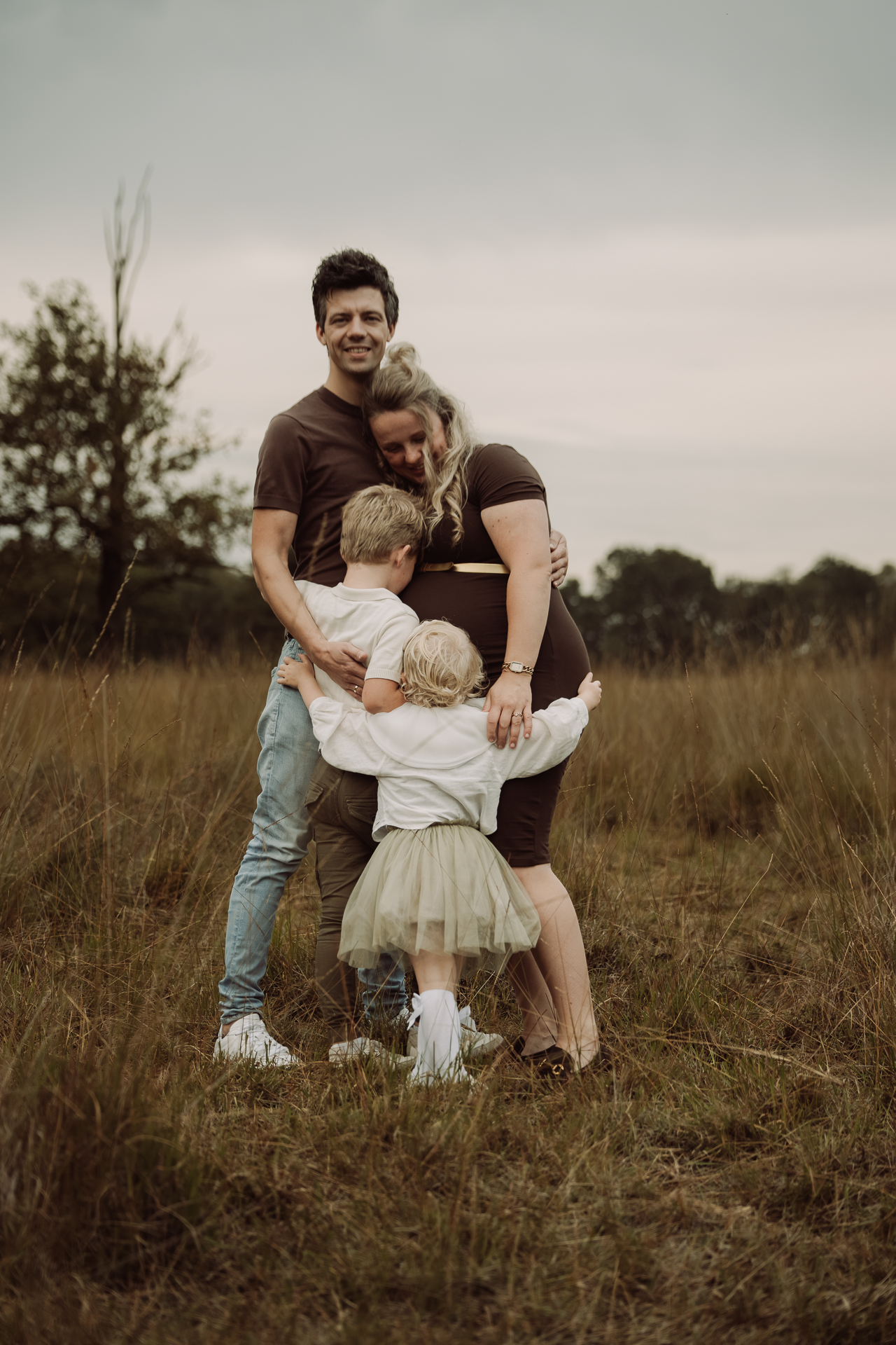Twee kinderen knuffelen hun ouders, terwijl hun zwangere moeder tegen haar partner leunt