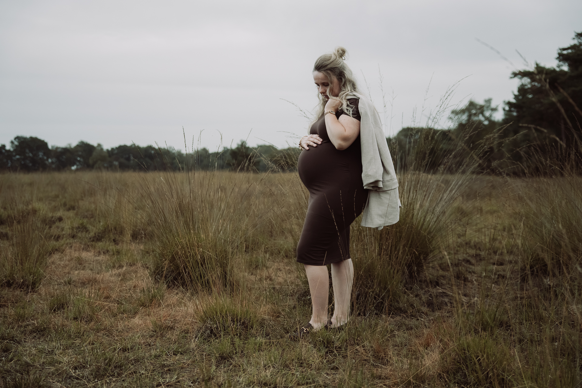 Zwangere vrouw met een vest over haar schouders kijkt naar haar buik tussen het hoge gras